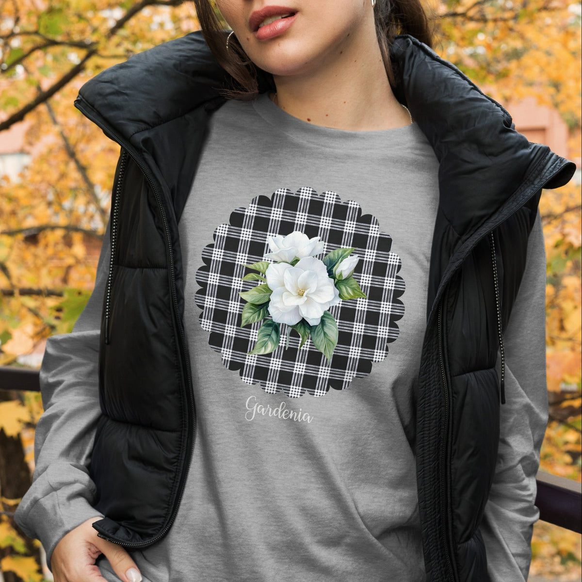 A woman wearing a heather gray long-sleeve t-shirt with a black and white gingham circle graphic featuring white flowers and green leaves, layered under a black puffer vest. The background shows a blurred outdoor scene with autumn trees.